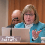 Joan Ballweg in front of a laptop and name plate, speaking into a microphone
