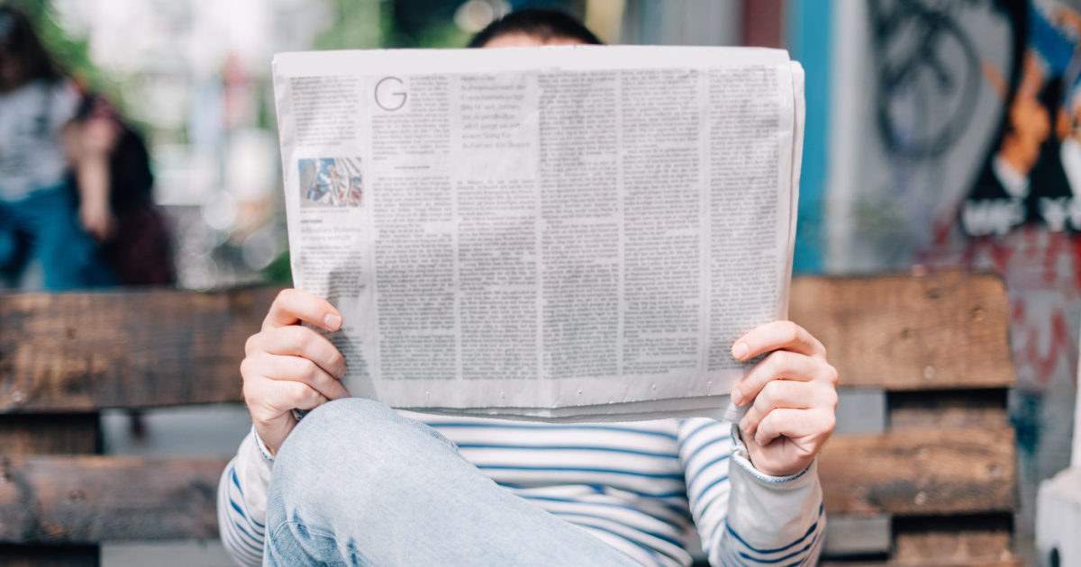 person sitting on a bench reading a newspaper that covers their face